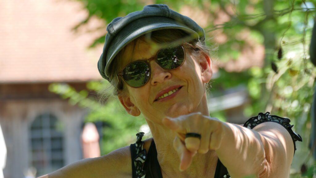 Portraitaufnahme ein Frau mit Hut und Sonnenbrille, die mit ausgestrecktem Zeigefinger in die Kamera zeigt.